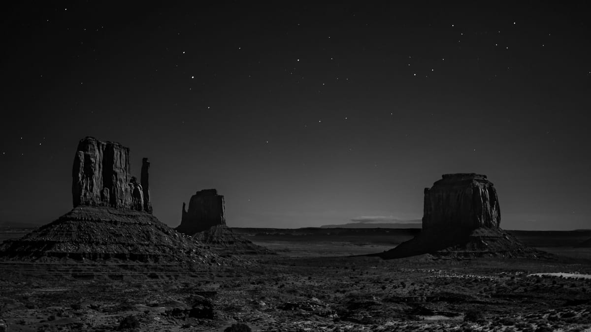 Desolate landscape of Monument Valley at night. Three silhouetted buttes under a starry sky evoke a serene and mysterious atmosphere.